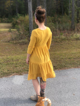 back view of short sleeve mustard with dot textured short dress that goes to knees. cinches at the small of the waist and flows out tiered on the bottom half